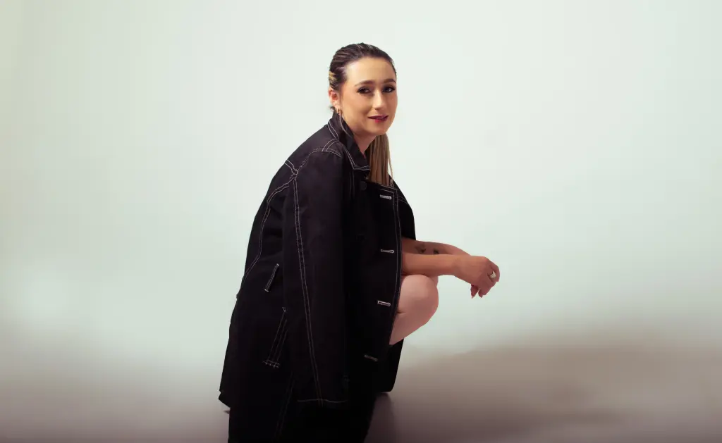 KateBlake poses in a black denim jacket, looking at the camera with her hair pulled back against a plain white background.