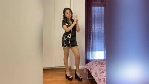 DalilaPearl — DalilaPearl stands in a black mini dress and high heels, posing with her hands together in a bedroom.