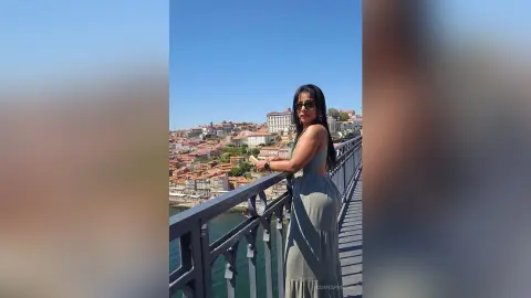 Stylish woman in a sage green maxi dress standing on a scenic bridge overlooking a coastal European city.