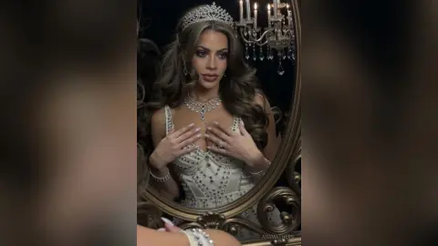 AnaMathers wears a silver tiara and a white beaded corset while posing in front of an ornate mirror.