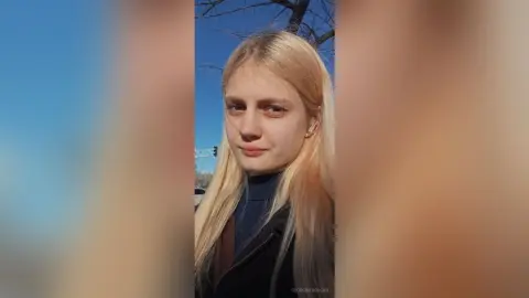 GeorgineAragan poses outdoors with long blonde hair and a dark blue turtleneck under a black jacket.