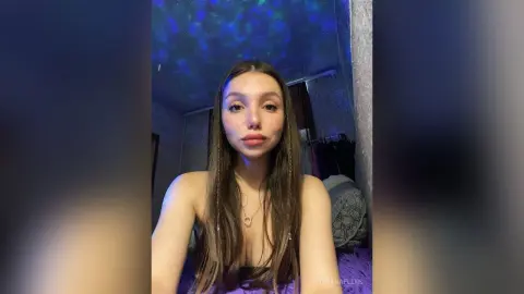 Young woman with long hair in a dark room under blue ambient lighting wearing a gold necklace.