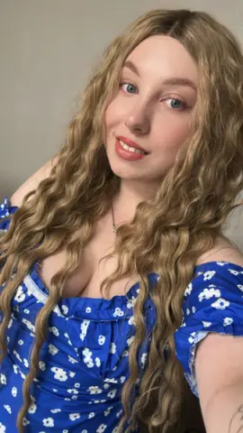 Smiling woman with long wavy blonde hair wearing a blue floral summer dress in soft natural lighting.