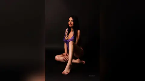NicolleSun poses in a purple lace bodysuit, sitting on the floor with her legs bent and hands resting on her thighs against a black background.