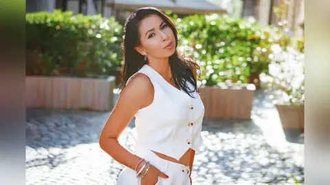 Smiling woman in a white sleeveless top posing in a sunny outdoor garden setting.