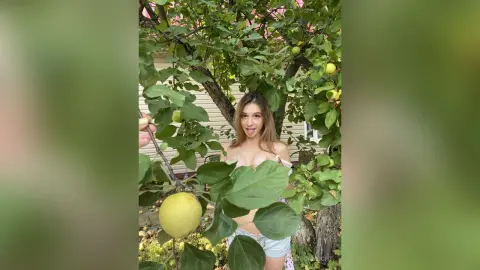 RitaMoonllight poses outdoors in a leafy tree, wearing light blue denim shorts and a white top that shows off her cleavage and bare shoulders.