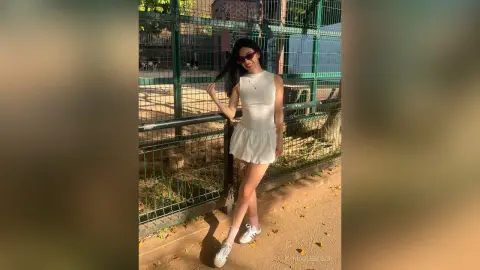 Young woman in a white sleeveless dress and sunglasses posing by a green fence in bright sunlight.