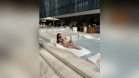 Woman in a bikini relaxing on a white lounge chair by a luxury rooftop swimming pool on a sunny day.