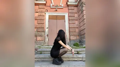 MarieJosePhilip, wearing a short black dress and black boots, crouches on stone steps in front of a large pink door.