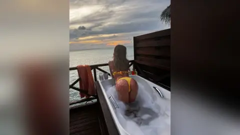 Woman in yellow bikini relaxing in an outdoor bathtub overlooking the ocean at sunset.