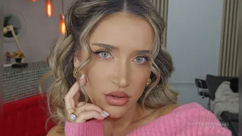 TiffanyAddams poses with her hands near her face, wearing a pink sweater and gold hoop earrings with her blonde hair styled in waves.