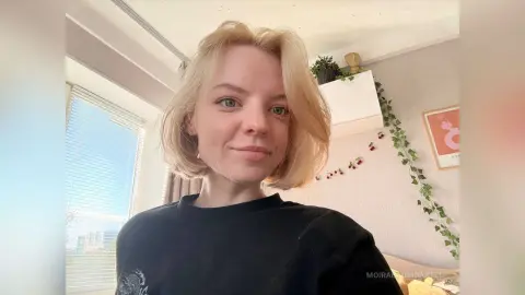 Smiling blonde woman in a black t-shirt sitting in a bright, cozy sunlit room.