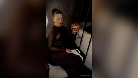 Elegant woman in a black long-sleeve outfit holding a cocktail in a moody, dimly lit lounge setting.