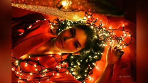 Elegant woman in a red dress lying amidst glowing fairy lights for a dreamy, festive holiday mood.