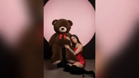 Young woman in a red satin dress hugging a giant brown teddy bear against a pink studio background.