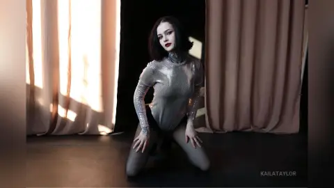Gothic woman in a shimmering silver bodysuit posing in a moody studio with draped curtains.