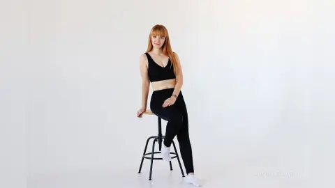 Redheaded woman in black yoga outfit sitting on a stool against a white background in a minimalist studio.