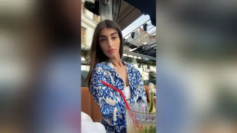 Young woman in a blue patterned shirt enjoying a refreshing drink at an outdoor cafe on a sunny day.