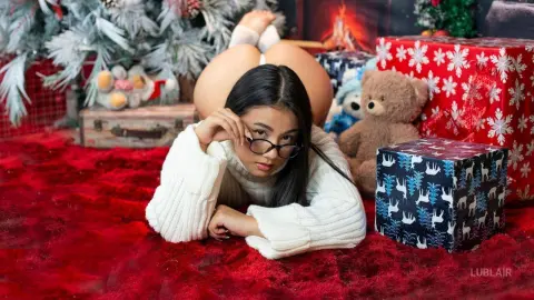 Young woman in a white sweater relaxing on a red rug near a teddy bear and Christmas gifts.