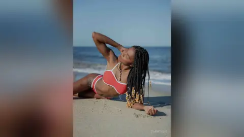 Smiling Black woman in a coral bikini relaxing on a sunny beach with ocean waves in the background.