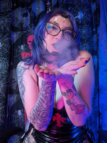 KimLennoxc with blue hair and glasses blows smoke from her hands while wearing a black outfit in front of a spiderweb backdrop.