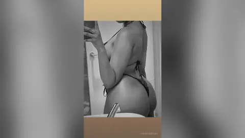 Black and white mirror selfie of a woman in a black bikini posing in a modern bathroom.