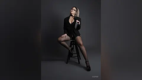 Elegant woman in a black blazer and sheer tights sitting on a stool against a dark studio background.