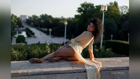 Woman in a flowing yellow dress sitting on a stone ledge in a lush green park during golden hour.