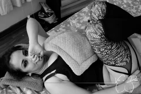 Victoria_Adams lies on a floral bed wearing black lingerie and fishnet stockings, looking at the camera with her hand near her face.