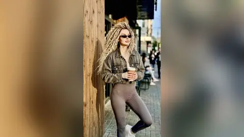 Blonde woman in denim jacket and leggings holding coffee while leaning against a wooden post on a sunny street.