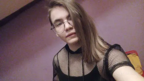 Young woman with glasses wearing a black mesh top in a dimly lit room with a moody aesthetic.