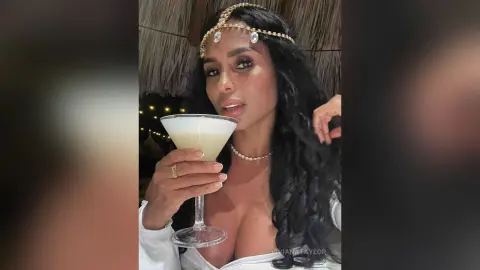 Glamorous woman in white outfit and gold headpiece sipping a cocktail in a tropical evening setting.