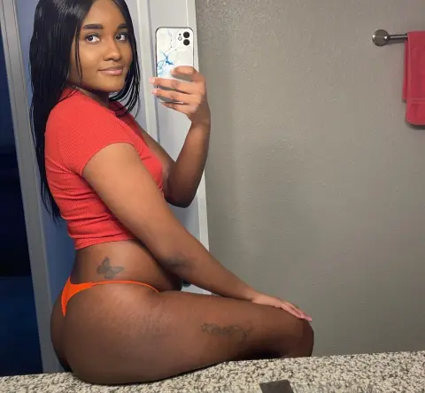 Smiling Black woman in a red crop top and orange briefs taking a mirror selfie in a modern bathroom.