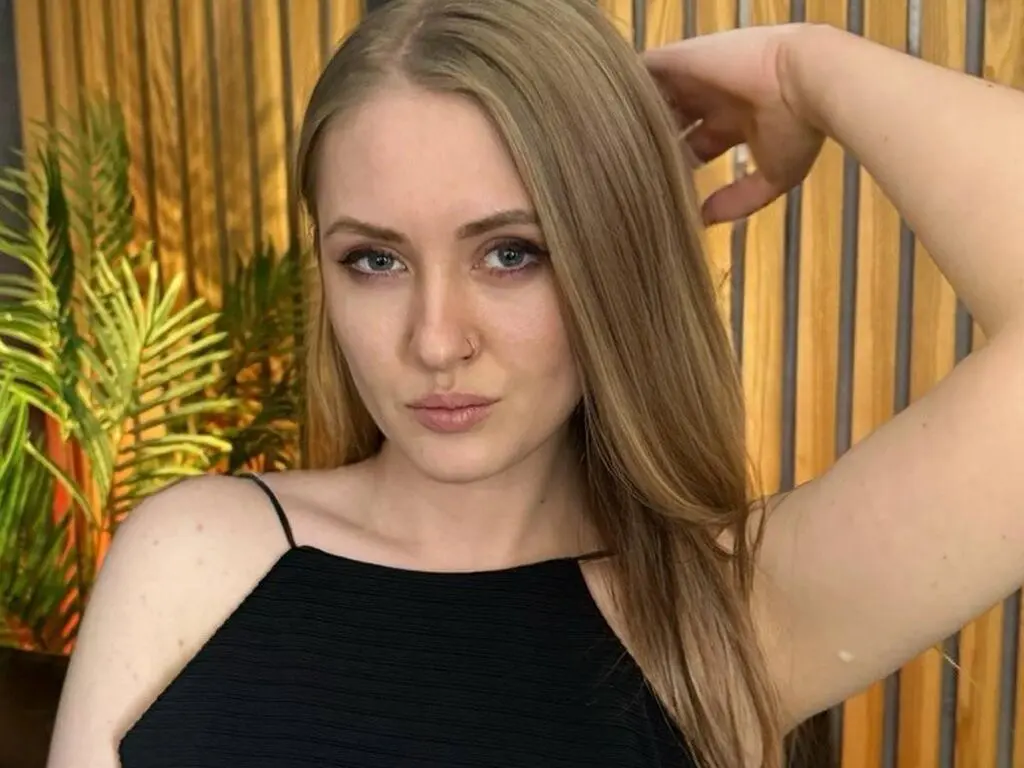 Blonde woman in a black tank top posing in a warm, sunlit room with tropical plants.