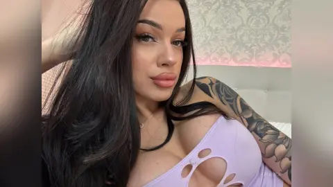 AriaVex poses with her hand in her long dark hair, wearing a light purple cut-out top that shows her cleavage.