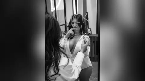 Moody black and white portrait of a woman in a white shirt looking into a mirror in a modern room.