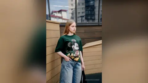 Young woman in a green Mickey Mouse graphic tee and baggy jeans standing outdoors by a modern building.
