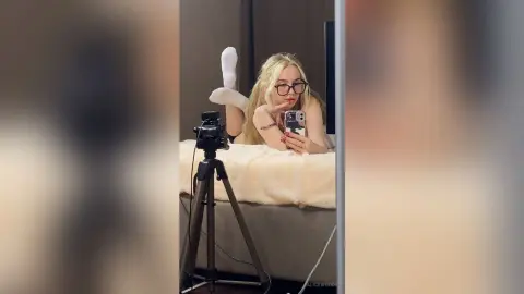 Blonde woman in glasses taking a cozy mirror selfie on a bed with a camera tripod in a modern bedroom.