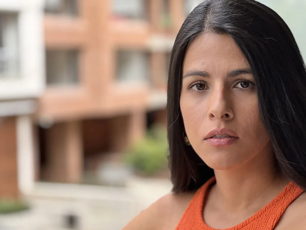 RaquelWilde looks into the camera with long dark hair and an orange top against a blurred building background.