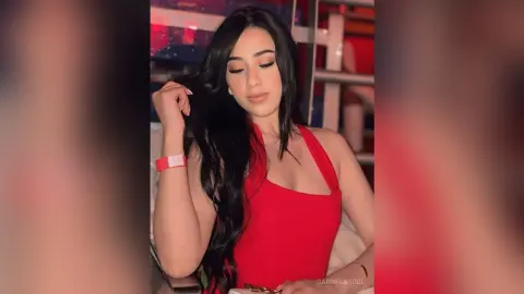 Young woman with long dark hair wearing a red halter top in a dimly lit indoor setting.