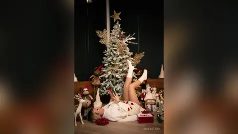 CamilaStarr lies on her back on the floor next to a white Christmas tree, wearing a white outfit and white socks with her legs raised in the air.