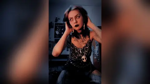 Goth woman with dark makeup and skull necklace posing in a moody, aesthetic bedroom setting.