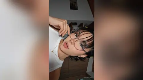 Young woman with short dark hair and glasses wearing a white tank top in a cozy indoor setting.