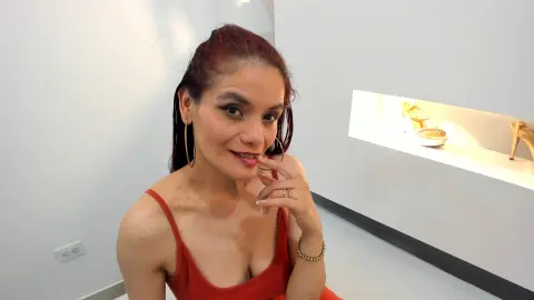 Smiling woman with red hair in a red tank top wearing gold hoop earrings in a bright modern room.
