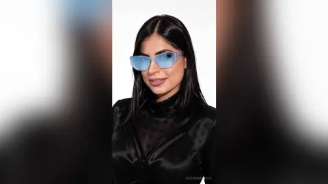 Stylish woman with long dark hair wearing blue tinted sunglasses and a black satin turtleneck in a studio setting.
