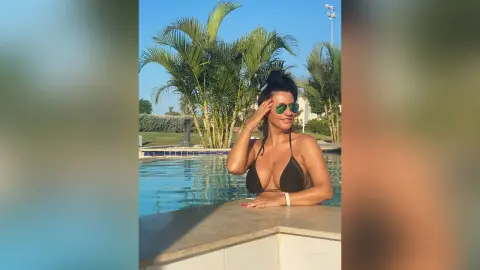 MissyJolie wears black bikini tops and sunglasses while leaning against the edge of a swimming pool surrounded by palm trees.