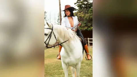 FlorenciaCopper wears a white shirt and brown cowboy hat while riding a white horse outdoors.