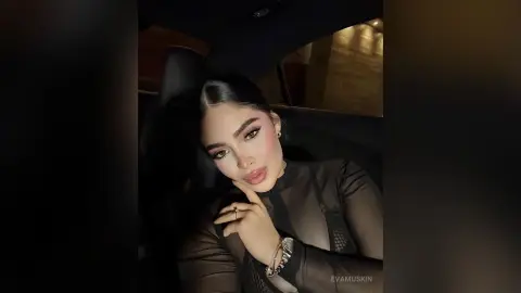 Glamorous woman in a sheer black top posing inside a car with elegant makeup and jewelry.