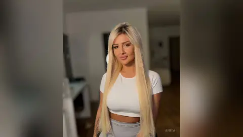 Anita poses in a white cropped t-shirt and grey leggings, letting her long blonde hair hang down over her shoulders.
