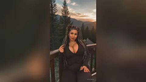 Curvy woman with long braids in a black bodysuit posing on a wooden balcony overlooking a misty mountain forest.